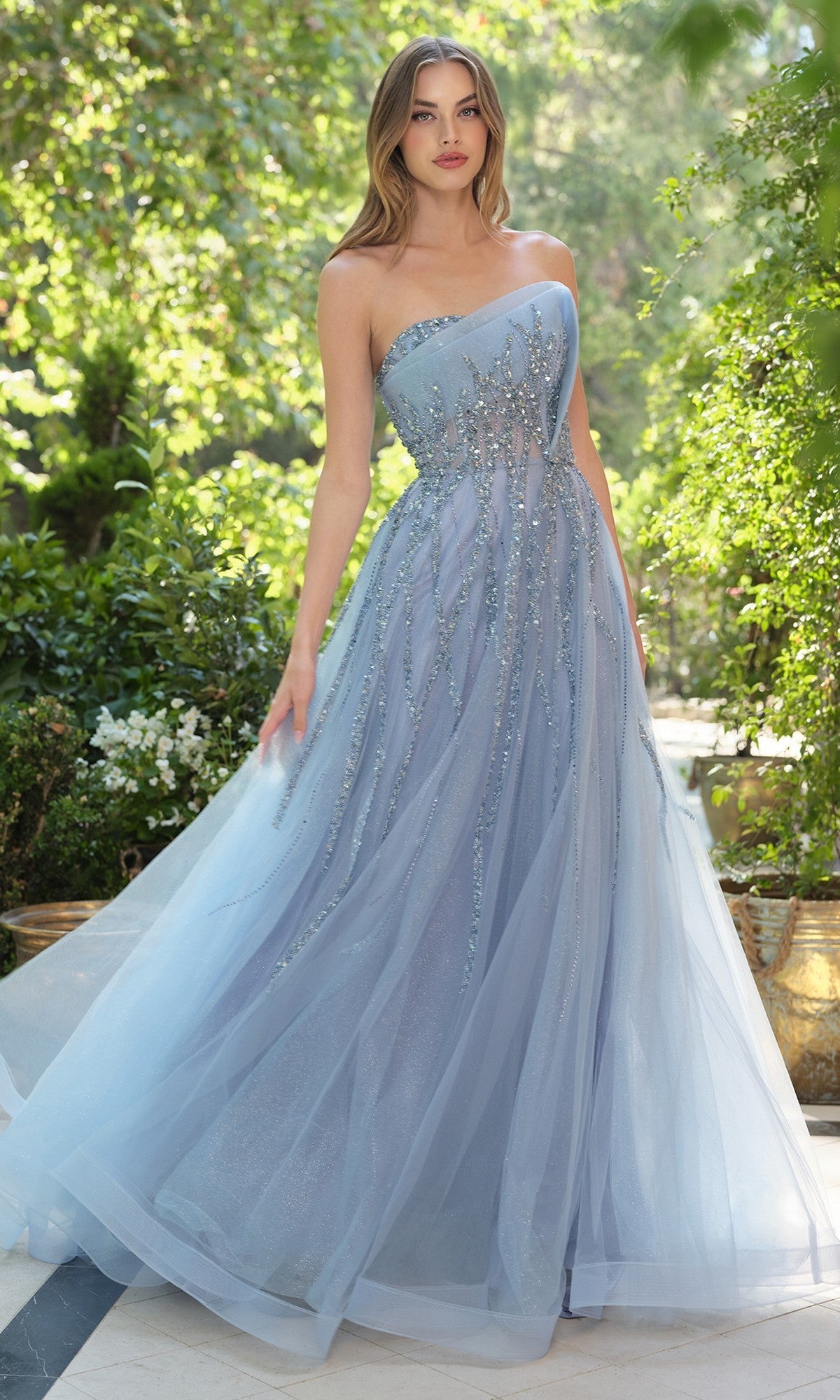 A woman stands outdoors in sunlight, wearing the Andrea and Leo A1491 long prom dress in smoky blue tulle with sparkling embellishments, surrounded by greenery.
